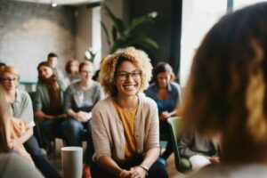 a sitting woman with curly hair and glasses smiles while group members sit behind her and their addiction specialist discusses benefits of cognitive-behavioral therapy 